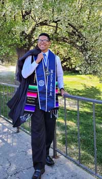 A graduate standing by the canal, gown over his shoulder, and colorful ropes and medals