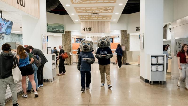 The Pack Place dining hall with multiple food stations labeled "FLAME," "DELI," and "STIR." University mascots, Alphie and Wolfie wear blue shirts and hold trays of food in the center of the image, surrounded by people walking and waiting in line. The scene captures a bustling, energetic atmosphere.