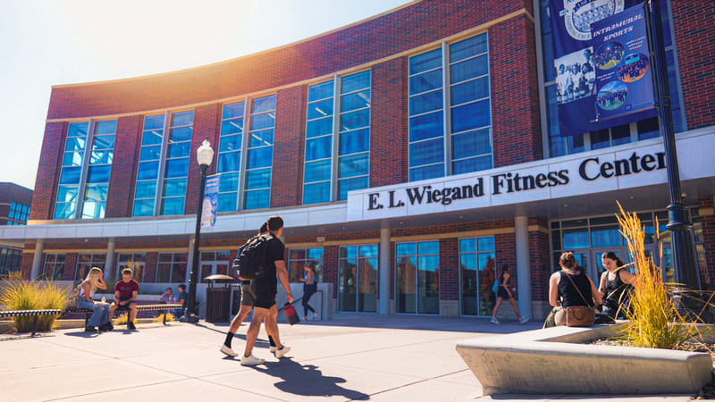 Students walk and gather outside the E. L. Wiegand Fitness Center on the University of Nevada, Reno campus on a sunny afternoon. The curved brick-and-glass facade of the recreation center is visible in the background, with campus banners, landscaping, and outdoor seating framing the entrance.