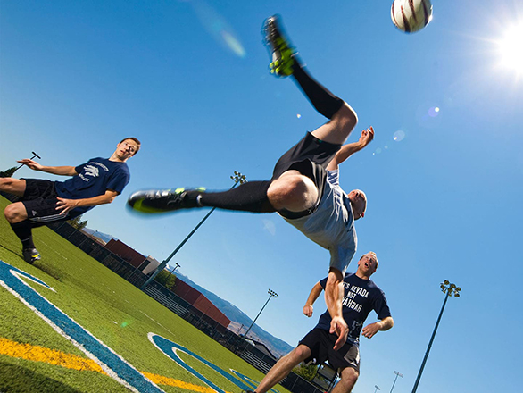 Three people playing soccer outside on a grassy field. One bicycle kicks the ball as two others watch.