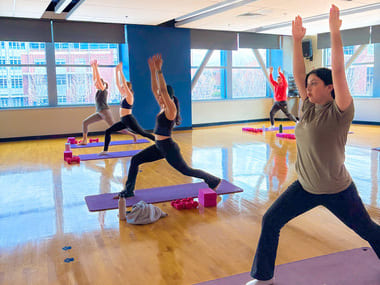 Participants practice yoga in a fitness studio inside the E. L. Wiegand Fitness Center. Students are positioned on yoga mats performing a standing lunge pose with their arms extended overhead, while natural light fills the room through large windows overlooking campus.