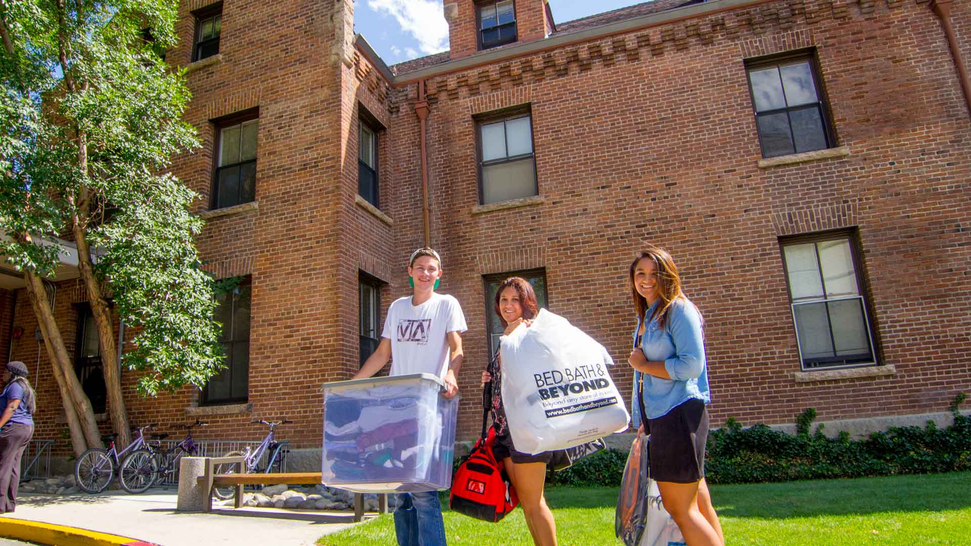 Three students stand outside Juniper Hall on move-in day, carring bins and bags with belongings