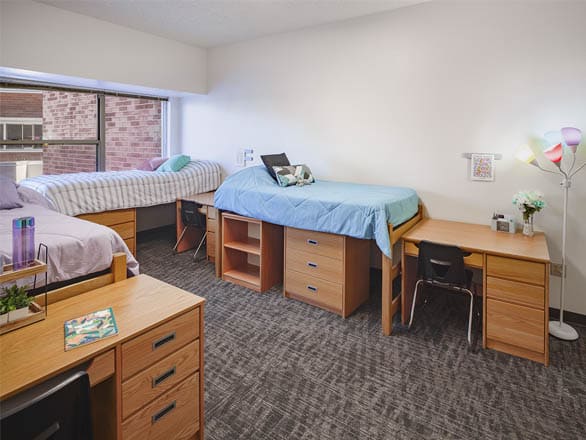 Spacious triple dorm room with three beds, personal desks, and storage space. Bright window light and cozy décor make it ideal for roommates.