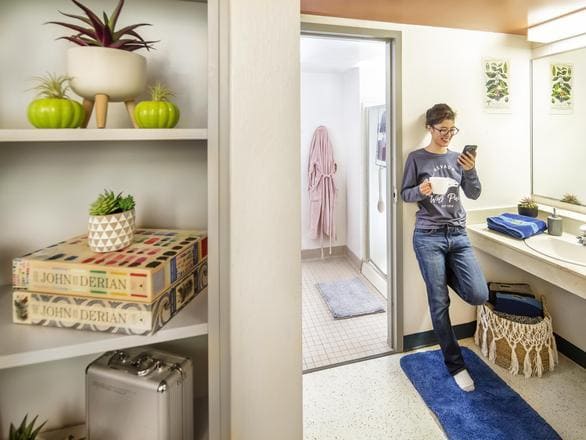 A person standing in a dorm bathroom in Canada Hall, holding a phone and a cup, with shelves displaying plants and books nearby.