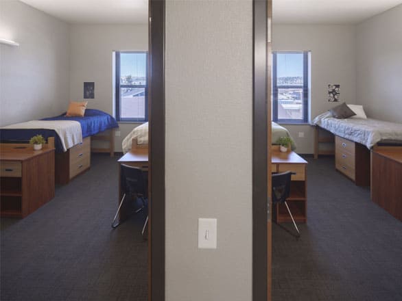 View of two adjoining double bedrooms separated by a central wall, each furnished with desks, drawers, and beds near bright windows.