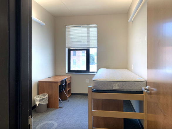 Single bedroom in Great Basin Hall featuring a lofted twin bed, wooden desk with chair, and large window providing natural light in a modern, minimalist student living space.