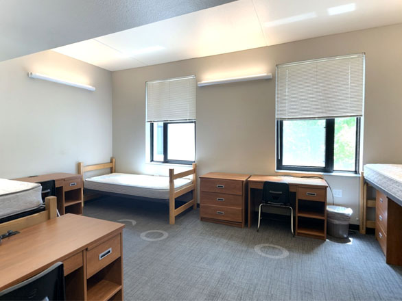 Triple bedroom in Great Basin Hall featuring three twin beds, each paired with a wooden desk and chair, in a spacious, naturally lit room designed for shared student living.