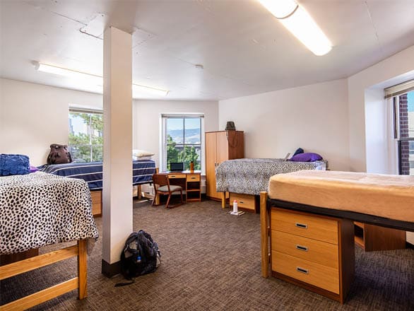  Large quad-style dorm room with four beds arranged around the space, two windows providing natural light, and shared desks and storage for a comfortable group living experience.