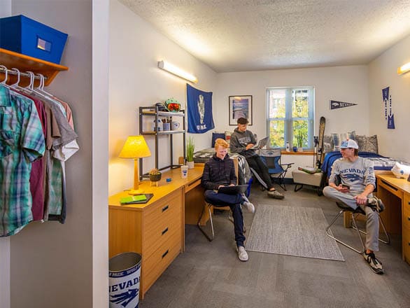 A bright, modern double bedroom in Nevada LLC Hall with two desks, chairs, and beds. Three students are sitting and relaxing, with University of Nevada banners and decor on the walls.