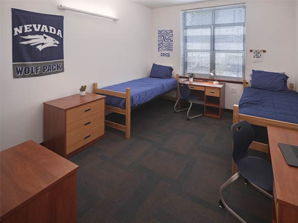 A bright, modern dorm room featuring two twin beds with navy bedding, wooden frames, and matching desks positioned under a large window. A Nevada Wolf Pack banner hangs on the wall, adding school spirit to the clean, minimalist space.
