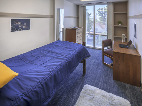 Cozy Sierra Hall single bedroom with one twin bed, a desk and chair, dresser, bookshelf, and a large window overlooking trees.