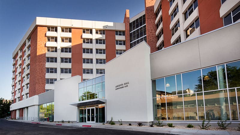Modern multi-story Argenta Hall with red brick and white accents, surrounded by trees and landscaped walkways under a clear blue sky.
