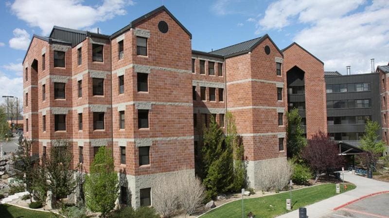 Canada Hall exterior at the University of Nevada, Reno, featuring a multi-story brick building with distinctive peaked roofs, surrounded by landscaped greenery and pathways under a partly cloudy sky.