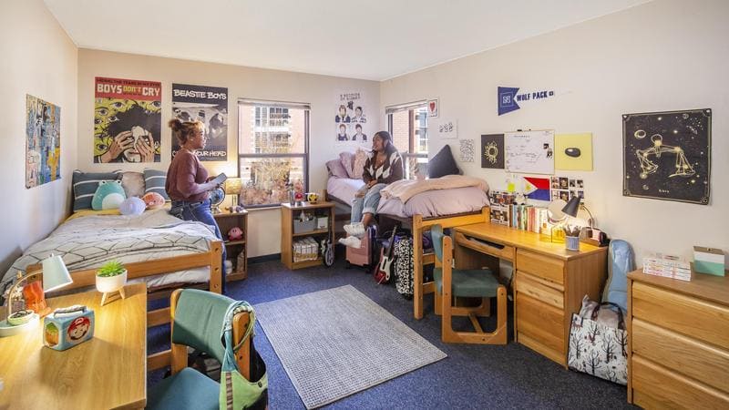 Two college students chatting in a bright, shared Canada Hall Hall room with beds, desks, and personal decorations.