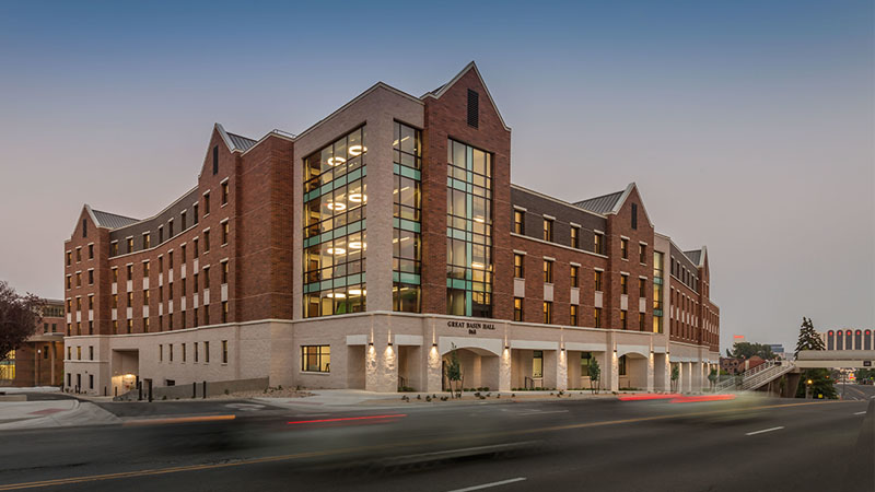 Exterior view of Great Basin Hall at the University of Nevada, Reno, showcasing its modern brick and glass architecture with tall windows and warm lighting at dusk.