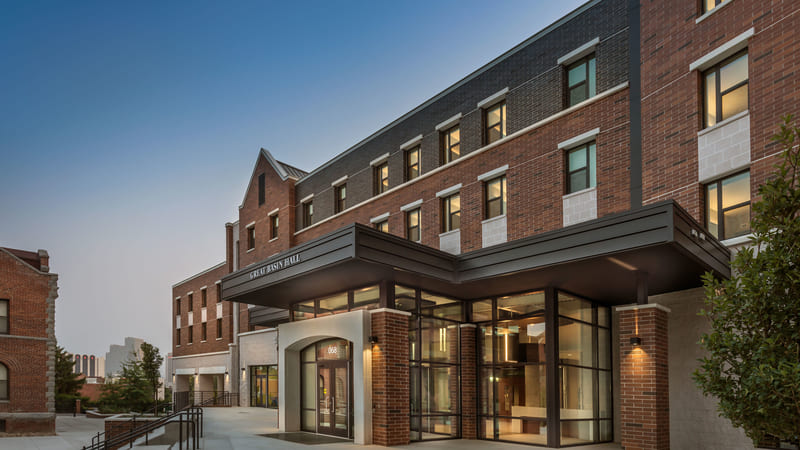 Exterior view of Great Basin Hall at dusk, featuring a modern brick façade with large glass windows and a prominent entrance canopy. The building has three stories with evenly spaced windows and a peaked roofline, creating a blend of contemporary and traditional architectural styles.
