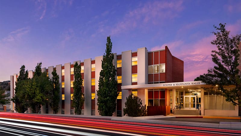 Exterior of Juniper Hall at the University of Nevada, Reno, featuring a modern mid-century design with vertical architectural lines, tall trees, and warm window lighting under a vibrant purple sunset sky.