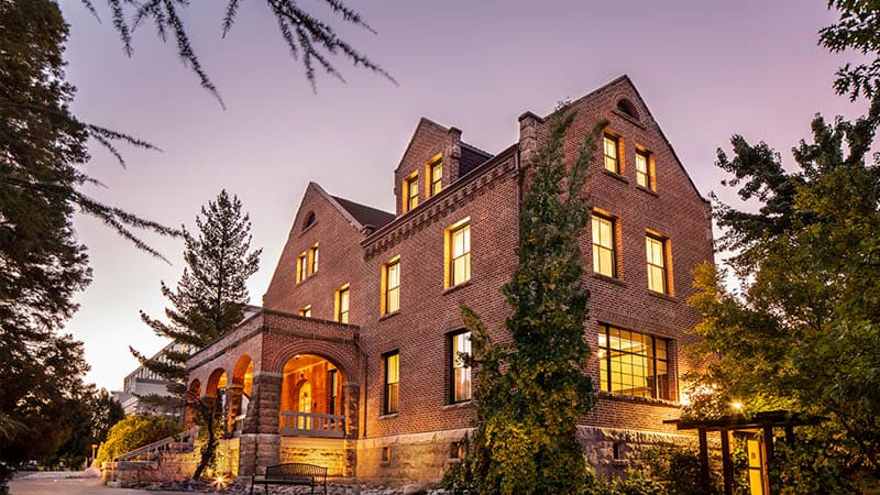Historic brick building of Manzanita Hall with warm lighting at dusk, surrounded by mature trees and landscaped pathways, showcasing its classic architectural charm.