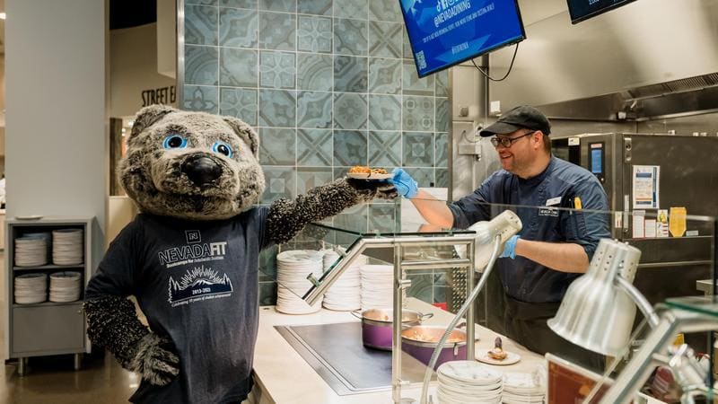 Alphie, the University of Nevada, Reno mascot, receives a plated meal from a dining staff member inside Pack Place dining hall, showcasing the welcoming atmosphere and variety of food options available to students.