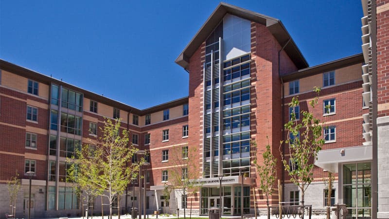 Nevada LLC Hall, a modern brick and glass residence building with a tall central entrance featuring vertical metal accents and large windows, surrounded by young trees under a clear blue sky.