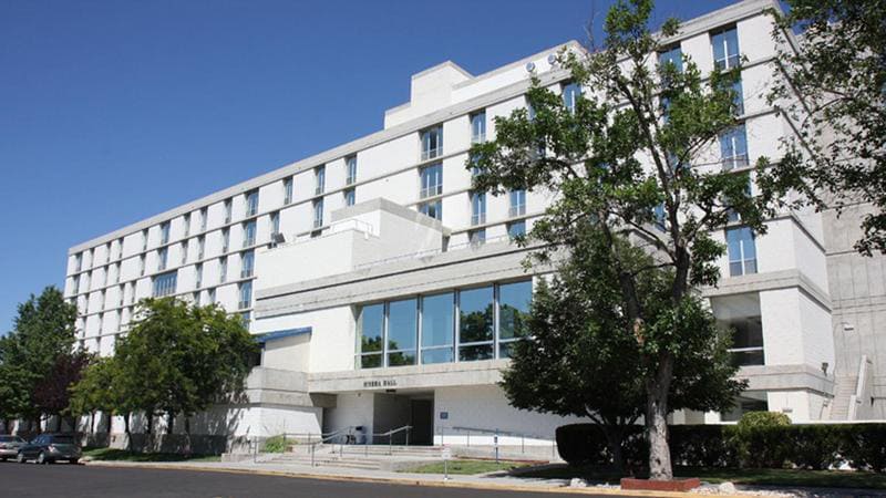 Exterior view of Sierra Hall, a multi-story white building with large windows on the lower level, surrounded by trees and a paved street in front.