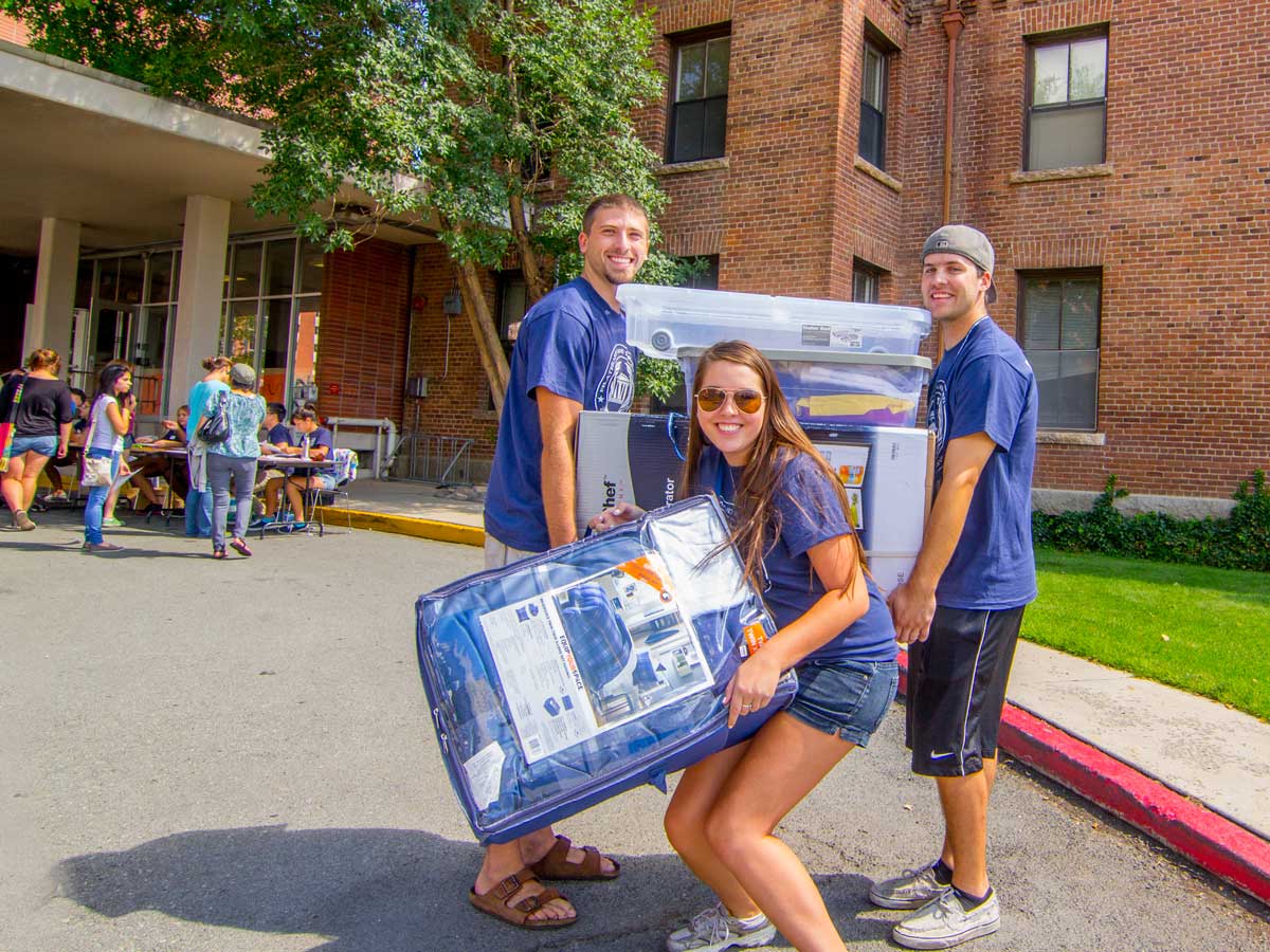 Three individuals wearing Wolf Pack gear pose outside a residence hall on move-in day
