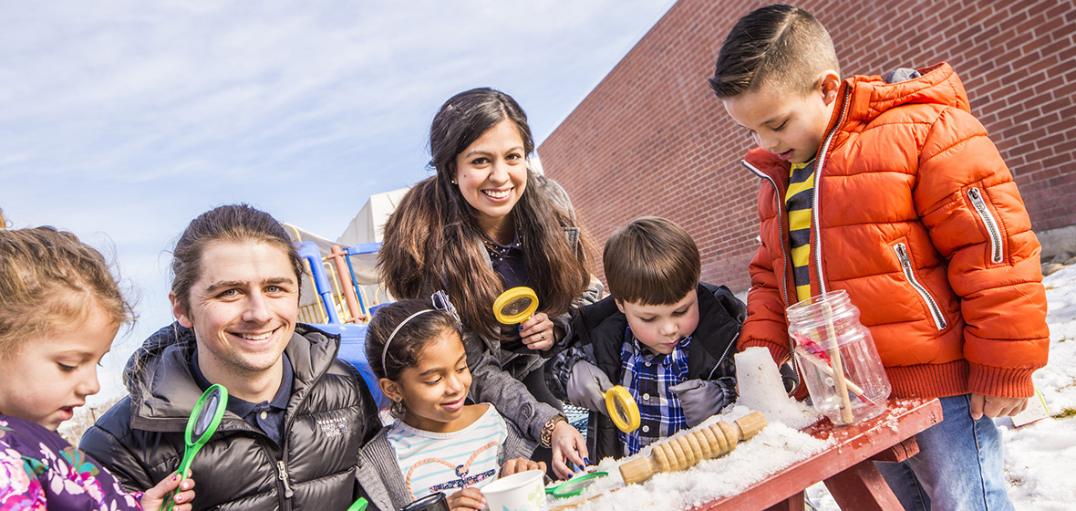 Three children playing with snow on a picnic table. They are accompanied by two smiling adults.