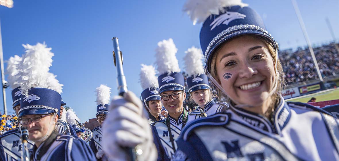 Wolf Pack Marching Band in full uniform marching out onto the field. A Piccolo player holds up her instrument and smiles into the camera.