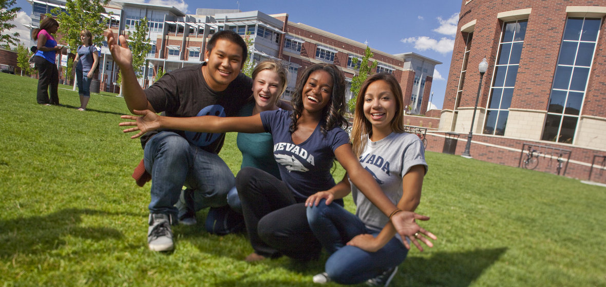 Four students in welcoming poses on the lawn in front of the Knowledge Center with the Student Union in the background