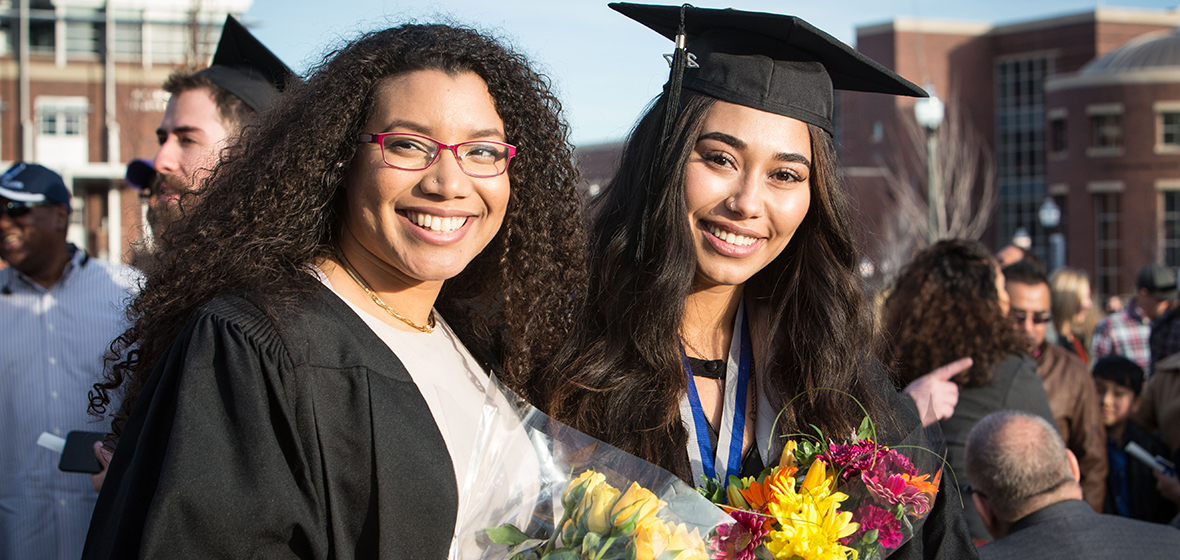 Two University graduates stand outside (with the Knowledge Center and Joe Crowley Student Union in the background), each holding flowers. 