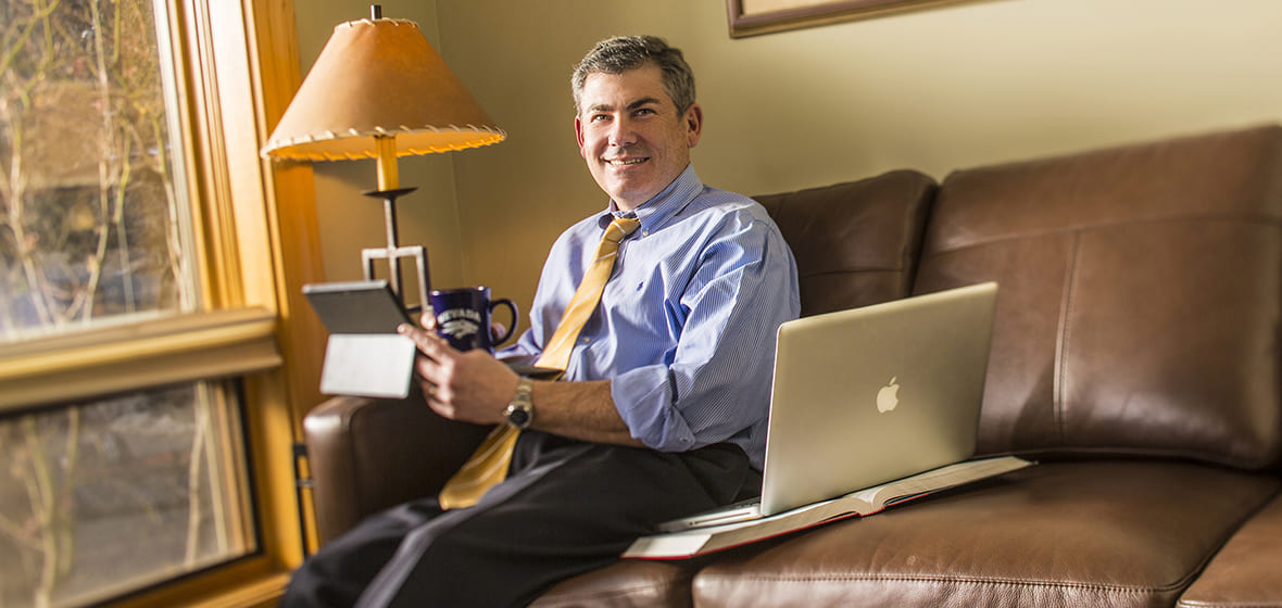A businessman sits on a leather couch with a book, a laptop, and a tablet, as well as a cup of coffee