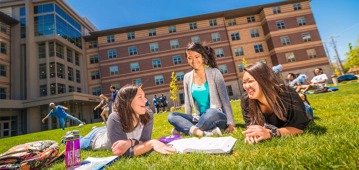 Three students lay or sit on the lawn in front of Peavine Hall on the University of Nevada, Reno campus