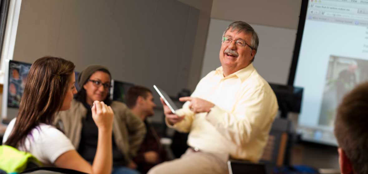 A man sits on a table in a classroom full of students. 