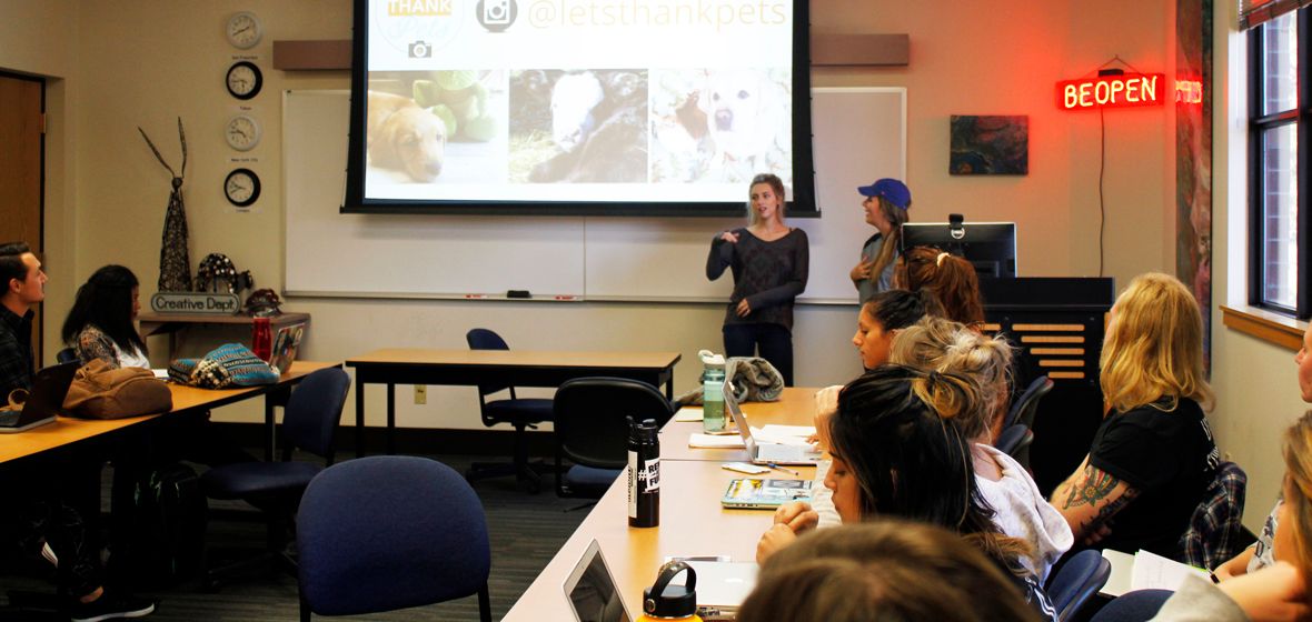 A group of students in a classroom. Two are standing at the front of the class. 