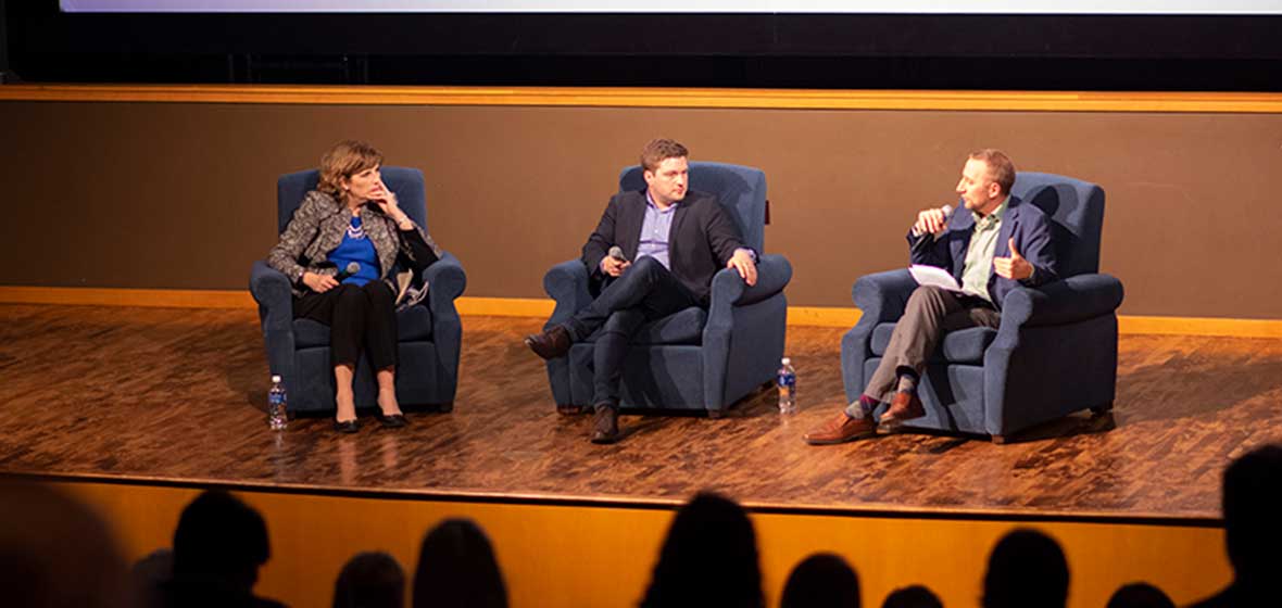 Three people sit in armchairs on a stage. 