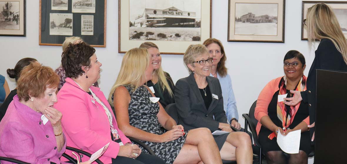 A small group of women are seated for an event inside the Reno Sparks Chamber of Commerce with Reno Mayor Hillary Shieve standing and talking to the group