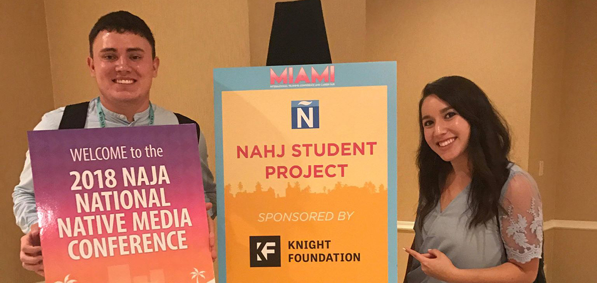 Male student holds sign, standing next to female student indoors.