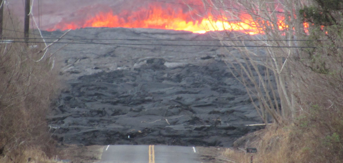 Lava flow in Kilauea swallows paved road with trees and a mailbox in the foreground and flames in the background