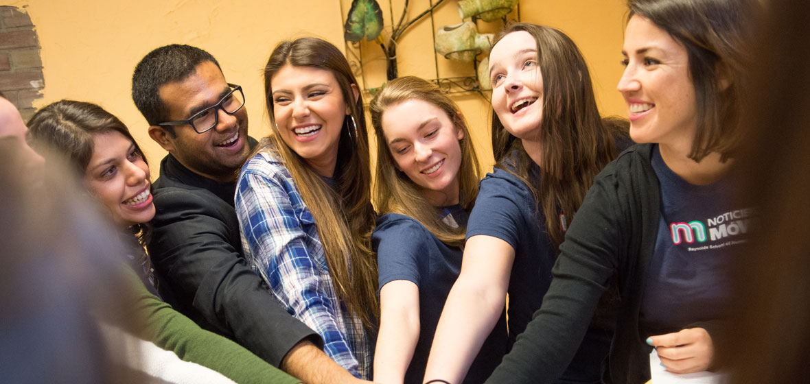 Five students and a professor place hands together in a circle indoors. 