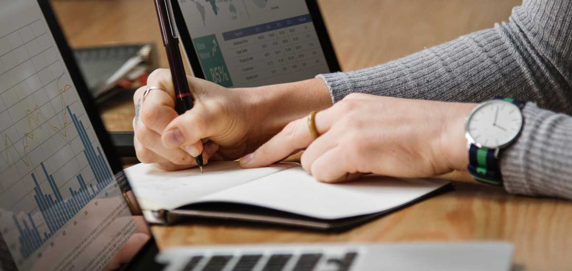 Hands can be seen writing on a notepad between two laptops 