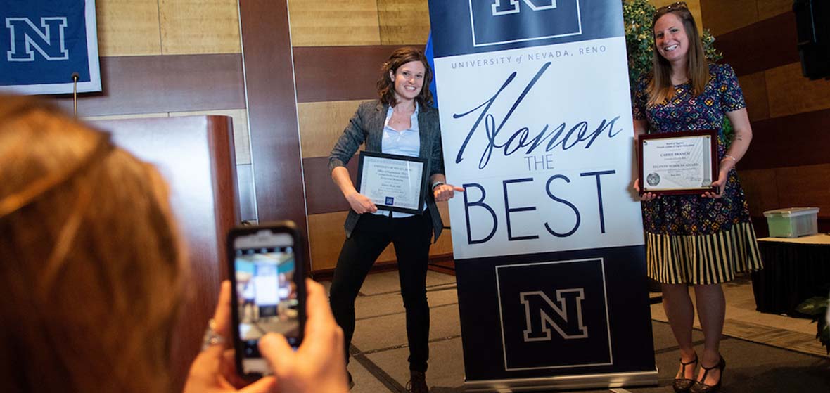 Two University of Nevada, Reno faculty members pose next to an Honor the Best banner while someone takes a picture of them with a cell phone