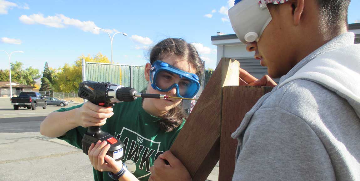 A girl drilling two wood pieces together as a boy holds it.