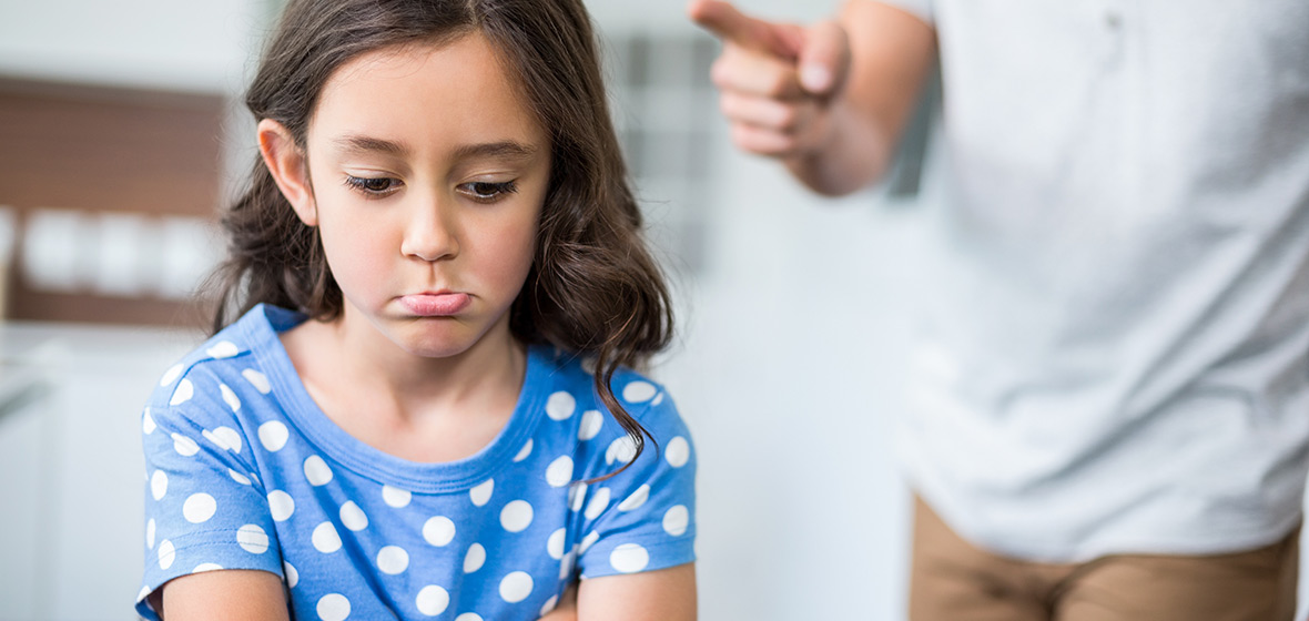 Little girl pouting with her bottom lip out while an adult in the background shakes a finger at her.