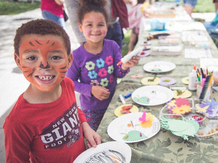Two children outside their with faces painted at a craft table with an adult.