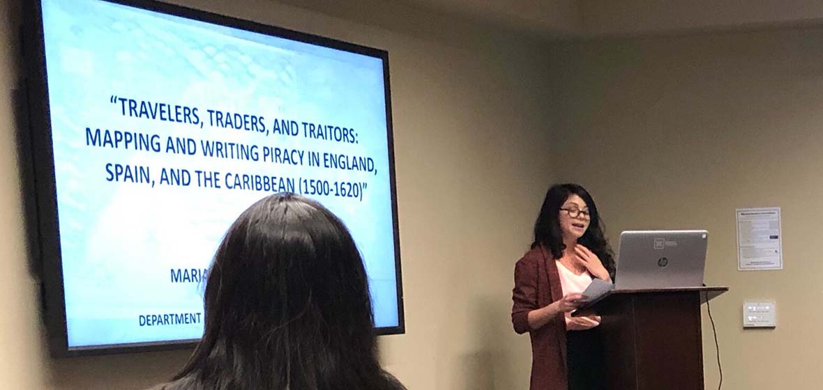 Mariana-Cecilia Velazquez behind a podium giving a presentation with a projector screen behind her and the back of an audience member's head.