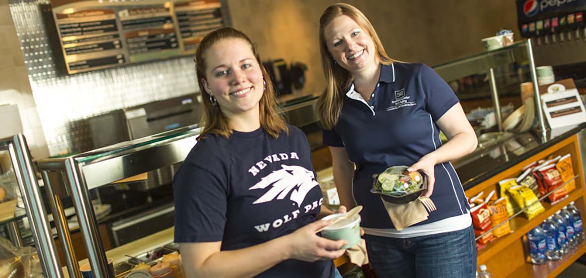 Two women in a deli stand and pose with food in hand