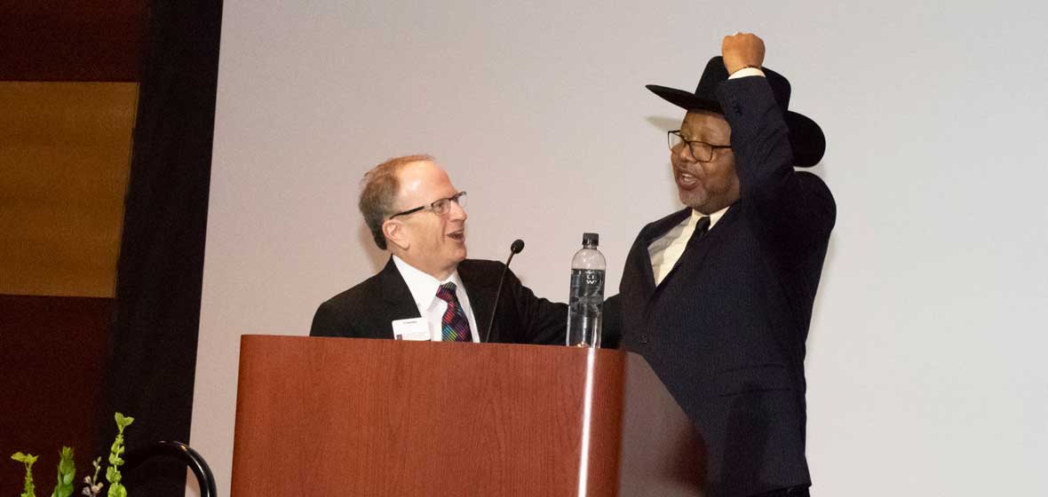 Two men on a stage, one of which is putting on a black cowboy hat. 