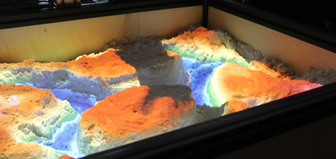 A student casts a shadow of their hand over the multicolored interactive sand table, which is designed to show changes in landscapes over time. 
