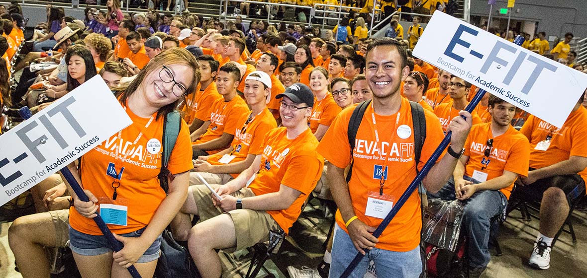 Students in orange shirts, some sitting and two standing, holding E-FIT signs as part of NevadaFIT's academic bootcamp kick-off ceremony in Lawlor Events Center. 