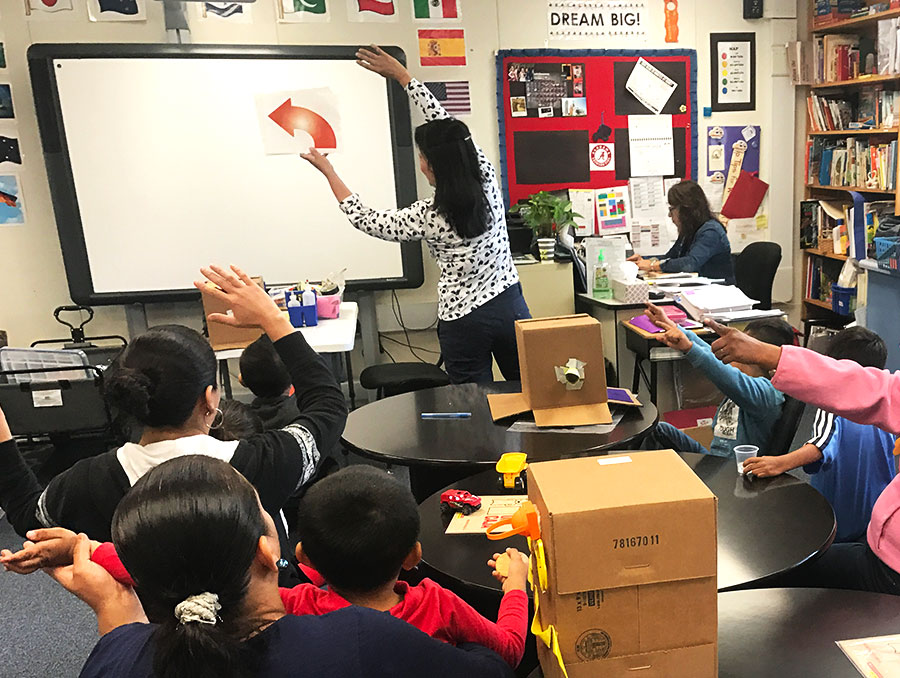 Lizeth holding an arrow sign and leaning in the direction of the arrow at the front of a classroom. Parents and children point in the arrow's direction.